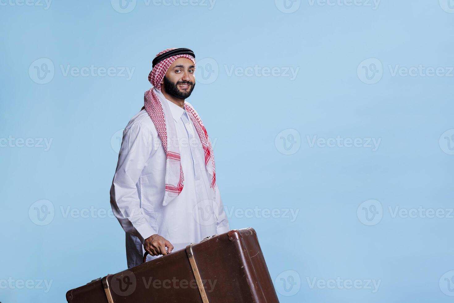 souriant musulman voyageur en portant ancien cuir valise et à la recherche à caméra avec de bonne humeur expression. content arabe habillé dans traditionnel islamique vêtements porter bagages studio portrait photo