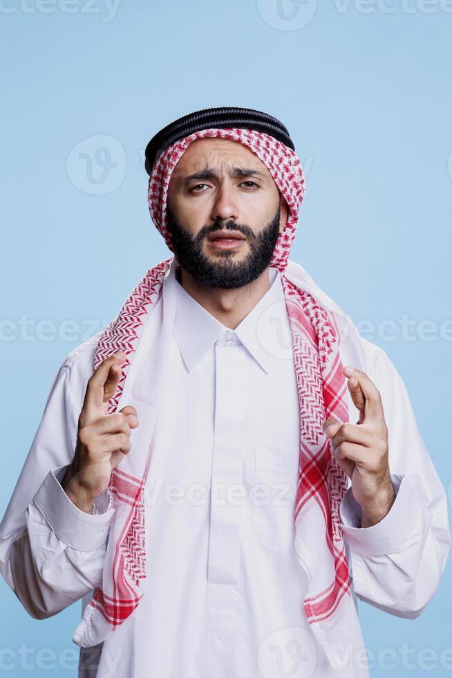 musulman homme habillé dans traditionnel vêtements fabrication souhait avec franchi les doigts et à la recherche à caméra avec optimiste expression. arabe portant thobe prier pour la chance avec superstitieux geste portrait photo