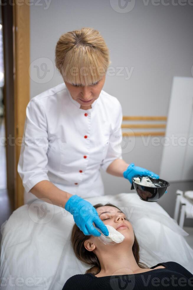 médecin cosmétologue ou dermatologue fabrication visage masque dans cosmétologie salon. professionnel esthéticien appliquant visage masque sur caucasien femme visage mensonge sur lit dans peignoir de bain. photo