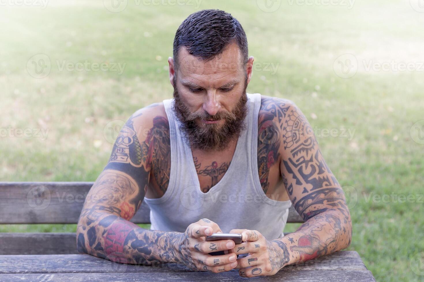 tatoué barbu homme écrit avec intelligent téléphone dans le parc photo