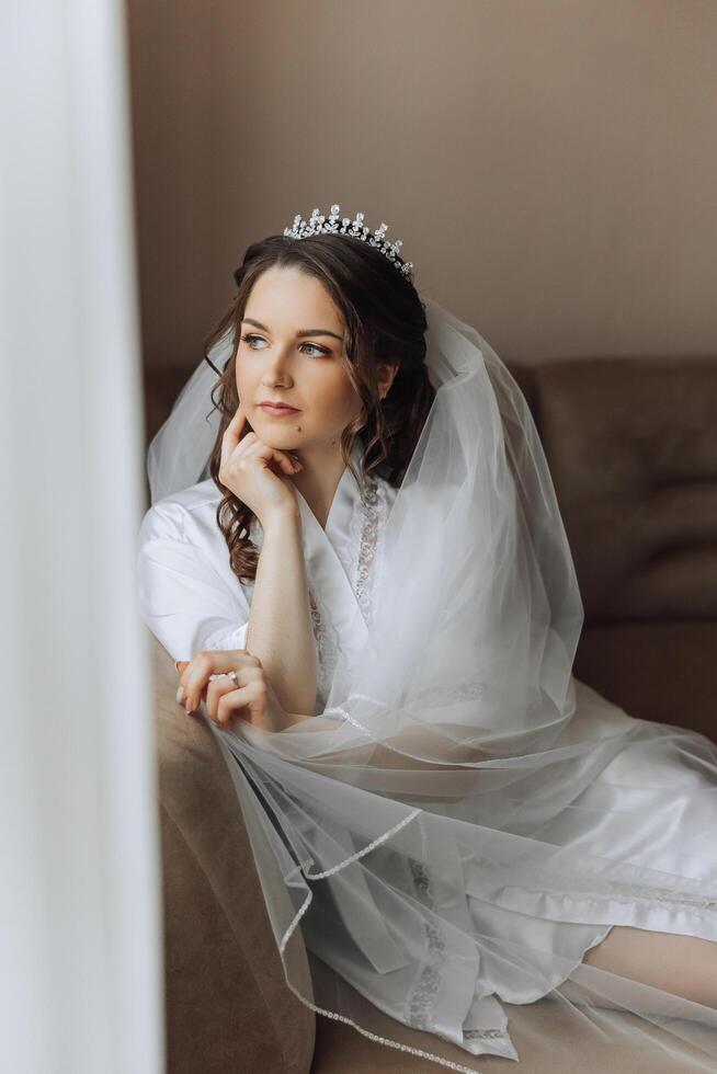 un incroyablement magnifique la mariée dans le Matin dans une blanc peignoir et une longue voile est assis sur le lit dans sa chambre à coucher. le la mariée pose dans le Matin avant le mariage cérémonie. photo