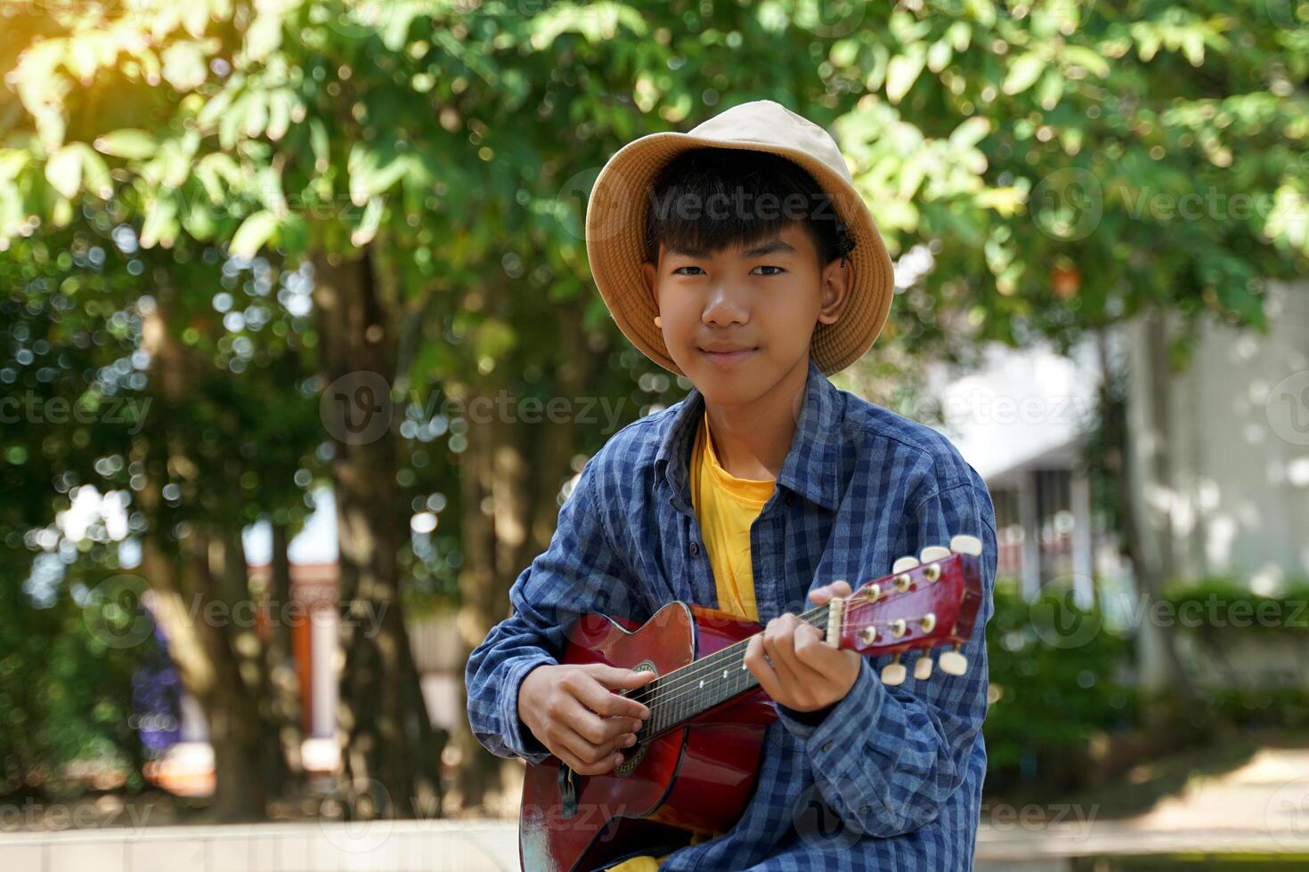 un asiatique garçon pièces le ukulélé pendant le sien gratuit temps dans le école Cour en dessous de le des arbres. doux et sélectif se concentrer. photo