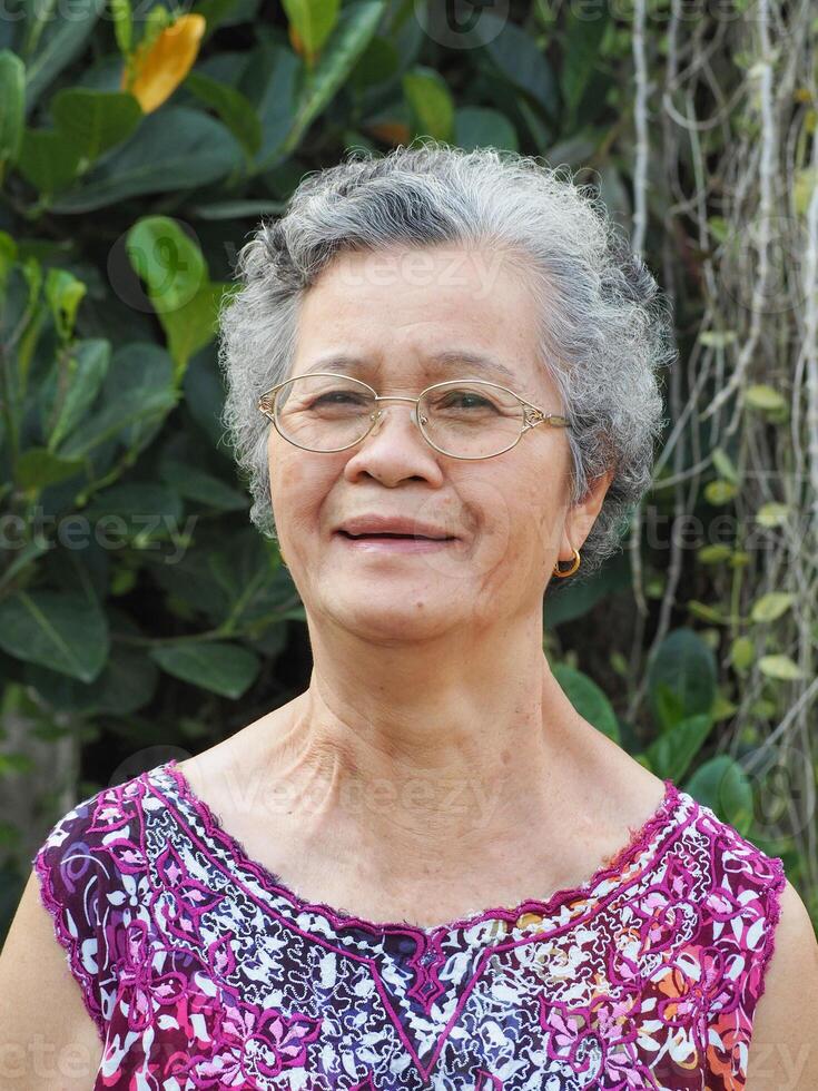 une Sénior asiatique femme avec court blanc cheveux souriant et à la recherche à le caméra tandis que permanent dans une jardin photo