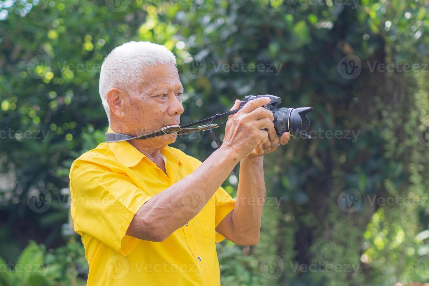 Sénior homme prise une photo par une numérique caméra tandis que permanent dans le parc. un personnes âgées asiatique homme porte une Jaune chemise être content lorsque en utilisant une caméra. concept de vieilli gens et la photographie