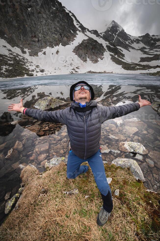 content actif de bonne humeur touristique dans Montagne des lunettes de soleil et une vers le bas veste, joyeux sourit expressivement se réjouit contre le toile de fond de une Montagne Lac haute dans le montagnes photo