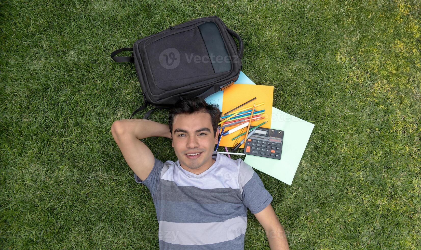 une Masculin étudiant mensonge sur le herbe avec une sac à dos et école Provisions photo