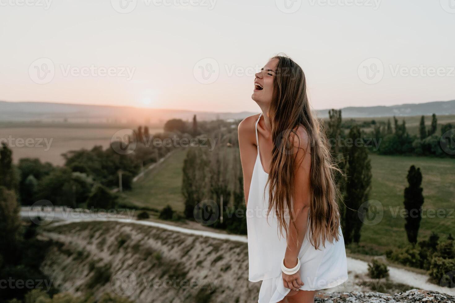 content femme permanent avec sa retour sur le le coucher du soleil dans la nature dans été avec ouvert mains. romantique magnifique la mariée dans blanc boho robe posant avec montagnes sur le coucher du soleil photo