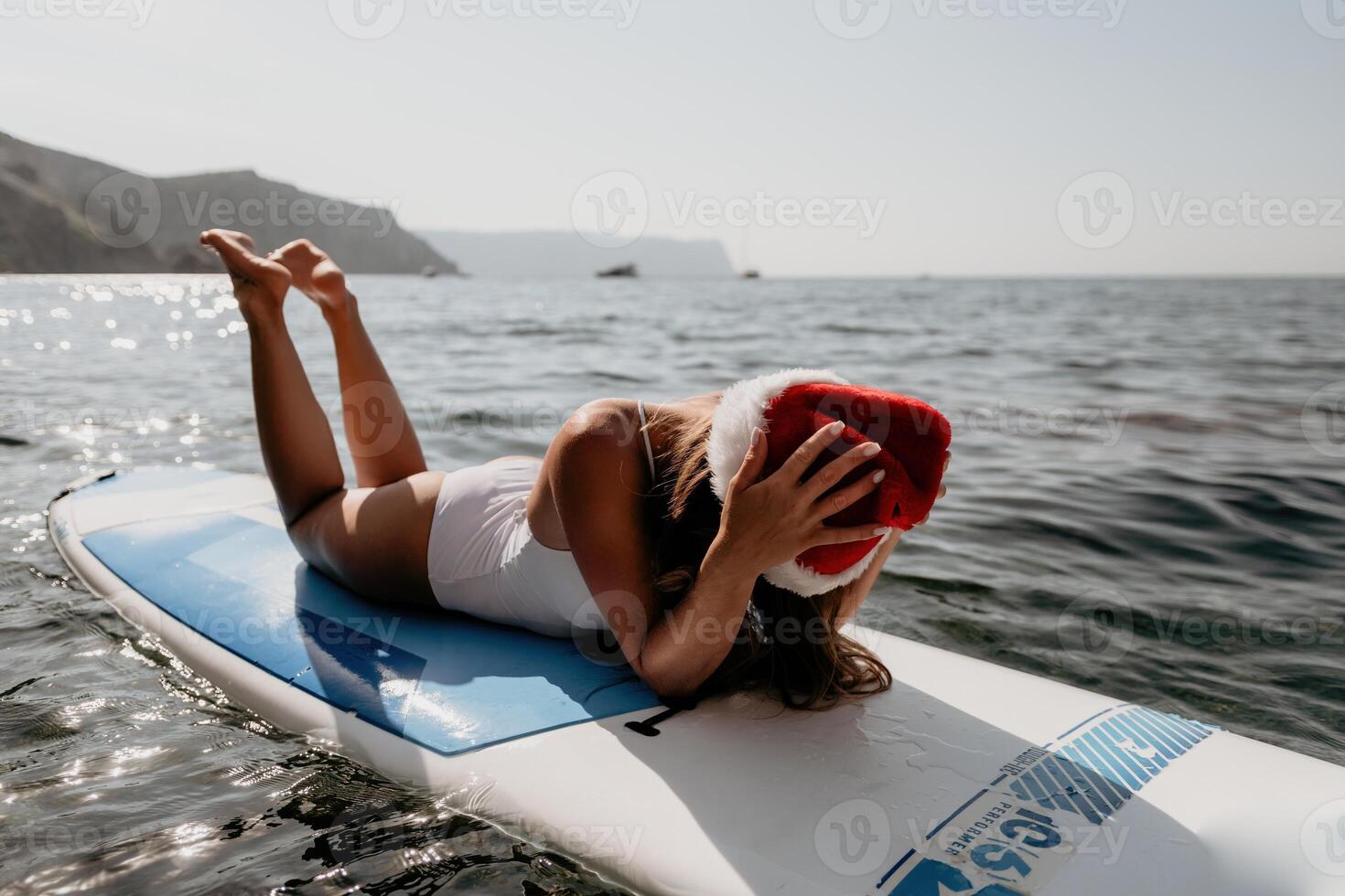 femme mer souper. proche en haut portrait de content Jeune caucasien femme avec longue cheveux dans Père Noël chapeau à la recherche à caméra et souriant. mignonne femme portrait dans une blanc bikini posant sur souper planche dans le mer photo