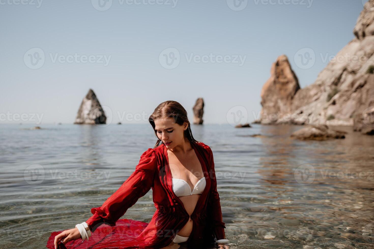 femme Voyage mer. content touristique dans rouge robe prendre plaisir prise image en plein air pour souvenirs. femme voyageur posant dans mer plage, entouré par volcanique montagnes, partage Voyage aventure périple photo