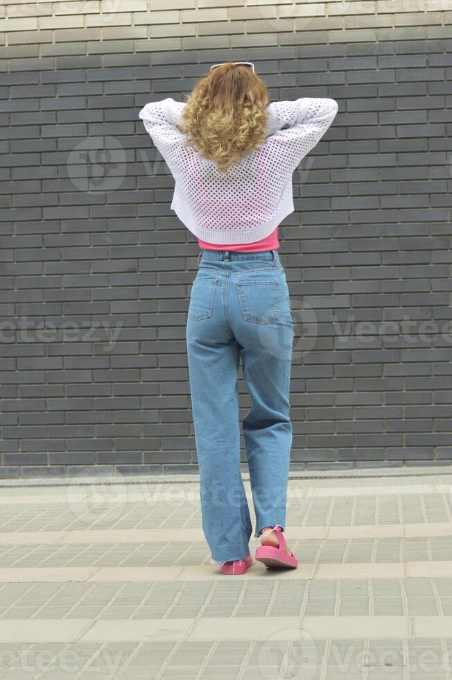 une blond fille dans jeans et une blanc chandail des stands avec sa retour à le caméra contre une gris brique mur. photo