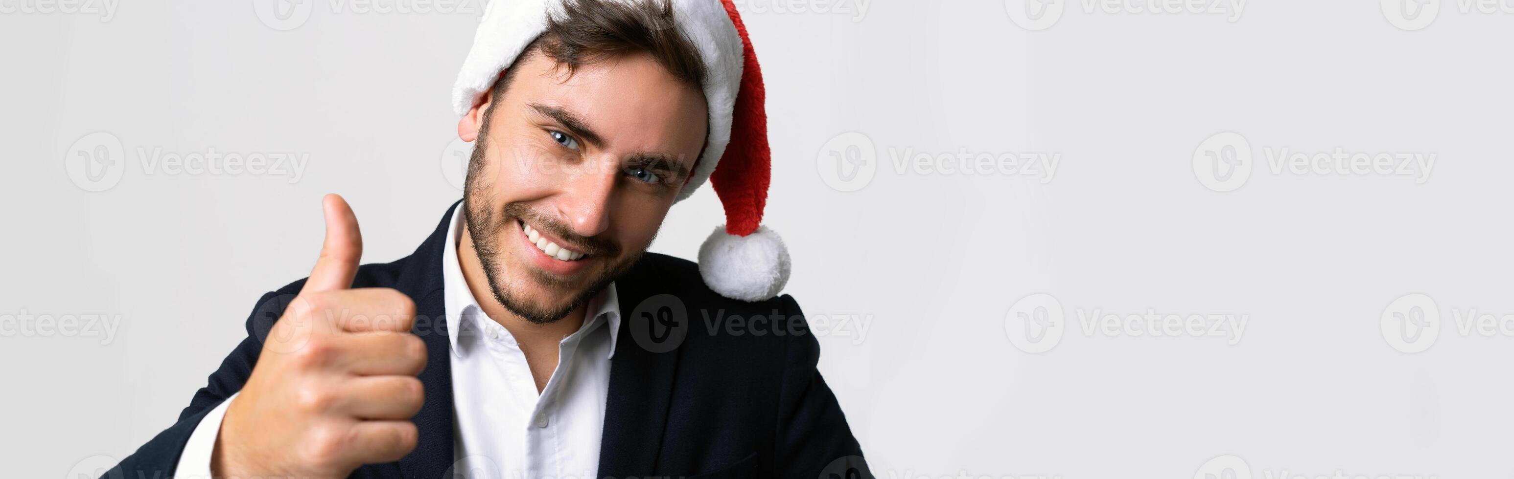 Jeune Beau caucasien gars dans affaires costume et Père Noël Chapeaux des stands sur blanc Contexte dans studio Smilie et montrant les pouces en haut. photo