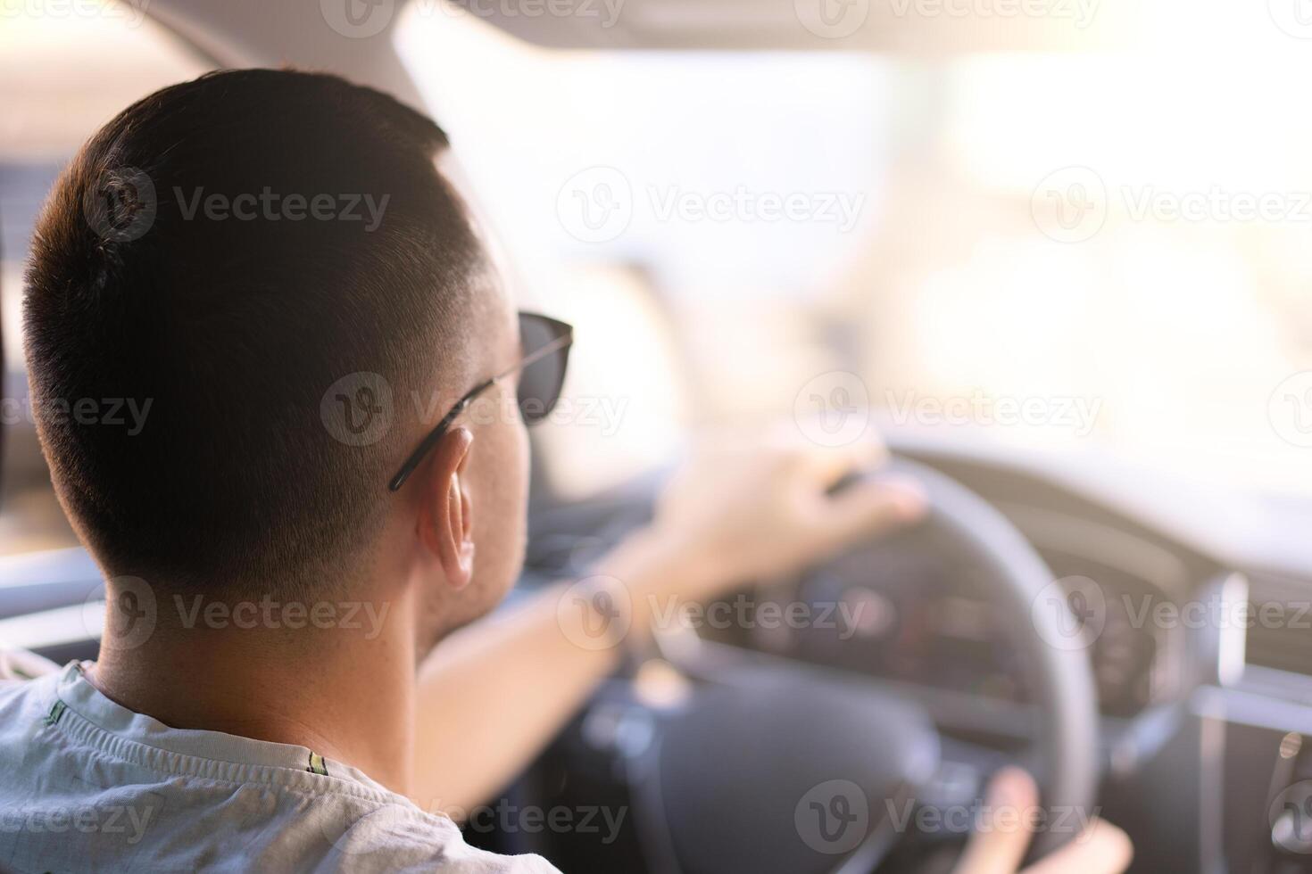 Jeune caucasien homme séance dans une voiture arrière vue en portant le pilotage roue en train de regarder le Traduction sur une brillant ensoleillé journée photo