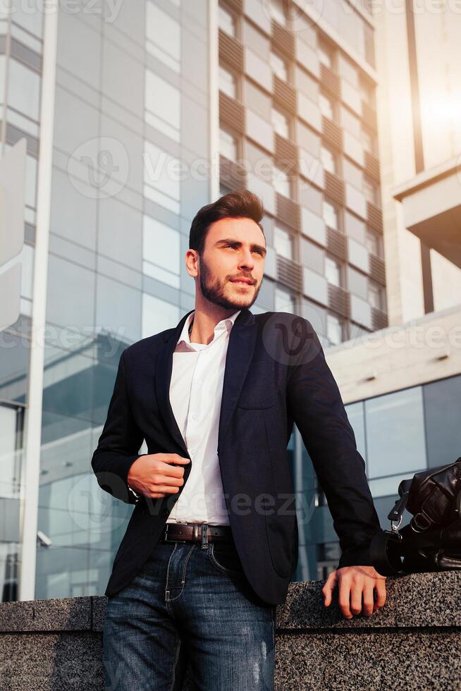 Beau Jeune homme d'affaire avec une barbe et dans une affaires costume permanent sur le rue contre le Contexte de le Bureau bâtiment suivant à une confortable élégant cuir sac. photo