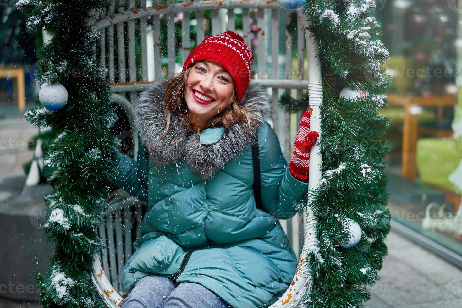 Jeune magnifique caucasien fille habillé dans une chaud hiver veste, tricoté chapeau et gants est assis sur Noël décoré balançoire Extérieur photo