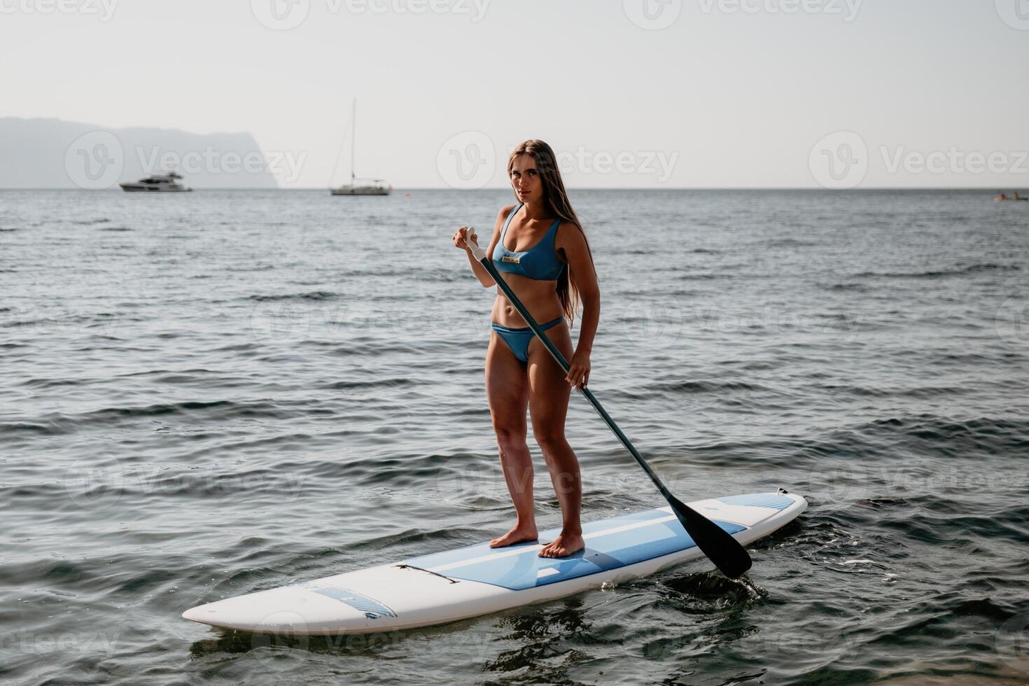 femme mer souper. proche en haut portrait de content Jeune caucasien femme avec longue cheveux à la recherche à caméra et souriant. mignonne femme portrait dans une bleu bikini posant sur souper planche dans le mer photo