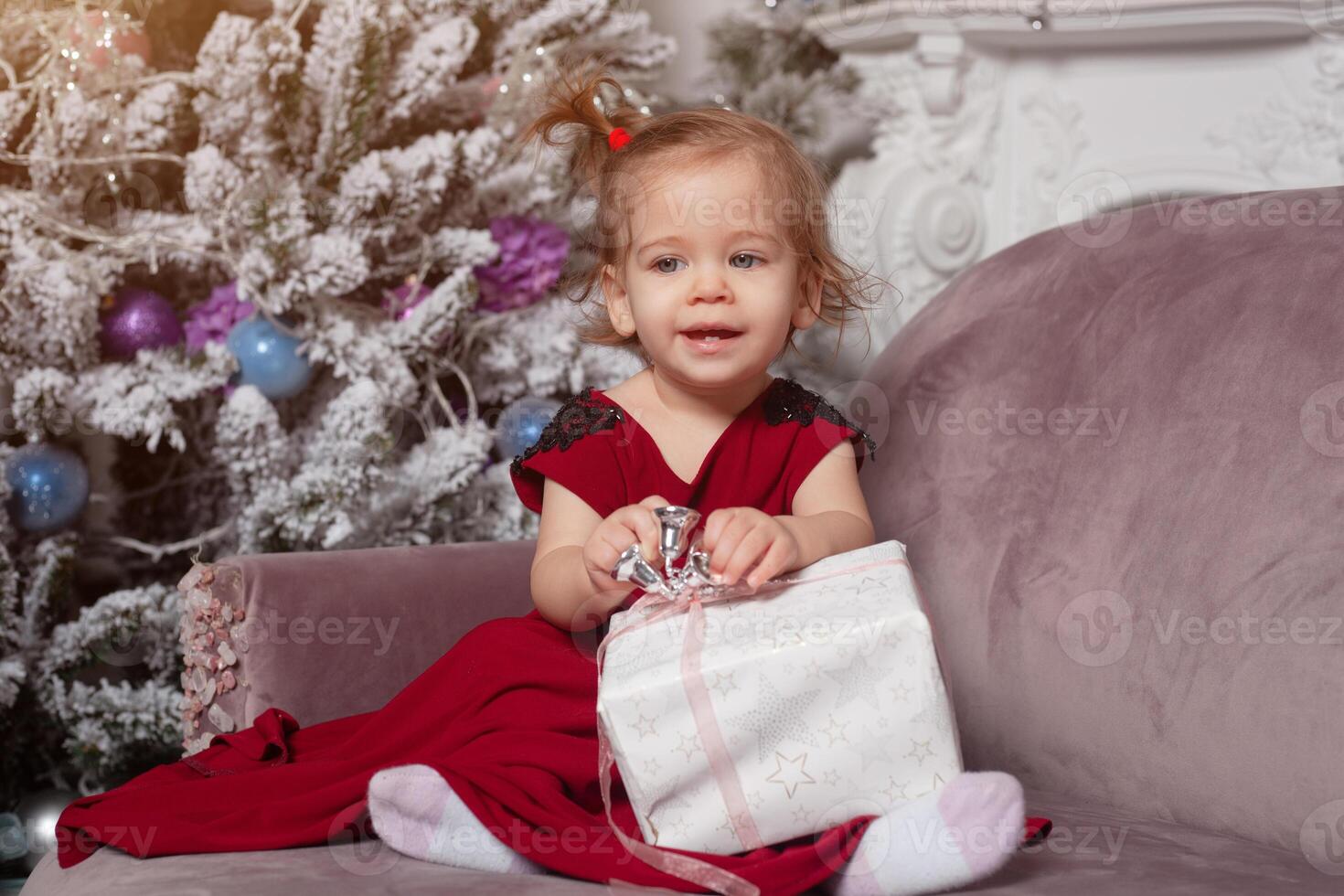 une magnifique mignonne peu fille habillé dans un élégant soir rouge robe est assis sur le canapé et ouvre une Nouveau années cadeau. avec Noël arbre sur le Contexte photo