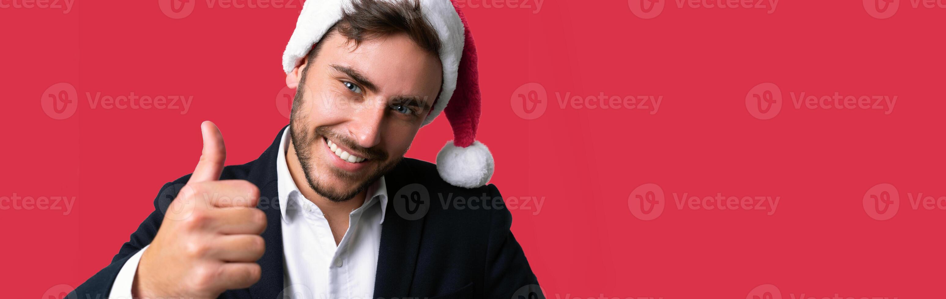 Jeune Beau caucasien gars dans affaires costume et Père Noël Chapeaux des stands sur rouge Contexte dans studio Smilie et montrant les pouces en haut. photo