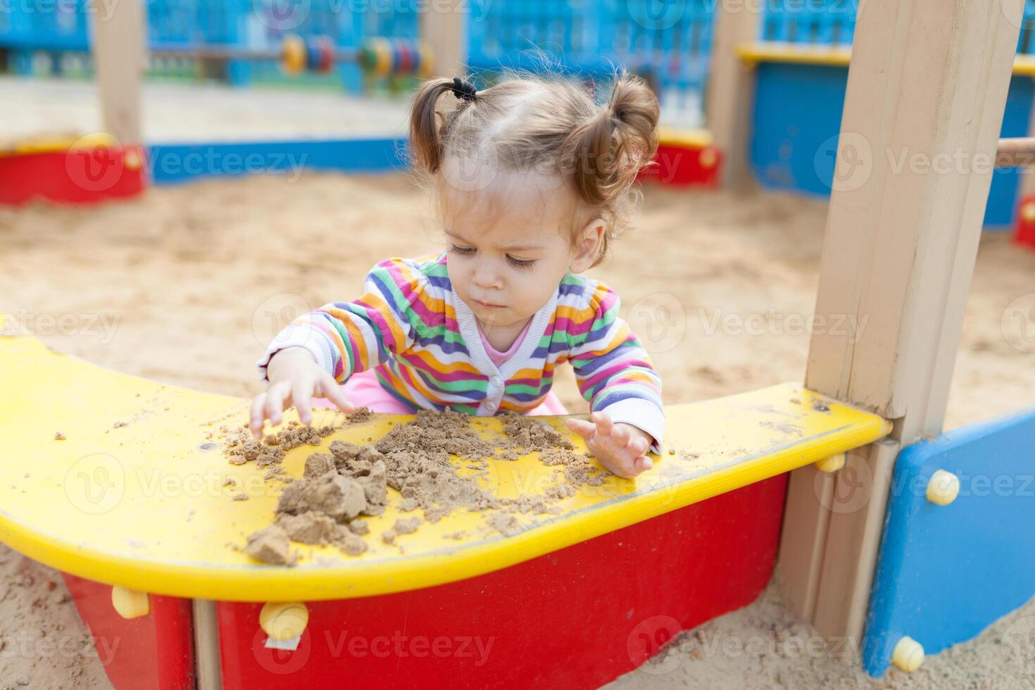 une peu fille avec deux queues est habillé dans une rayé coloré veste est en jouant dans le bac à sable sur le terrain de jeux photo