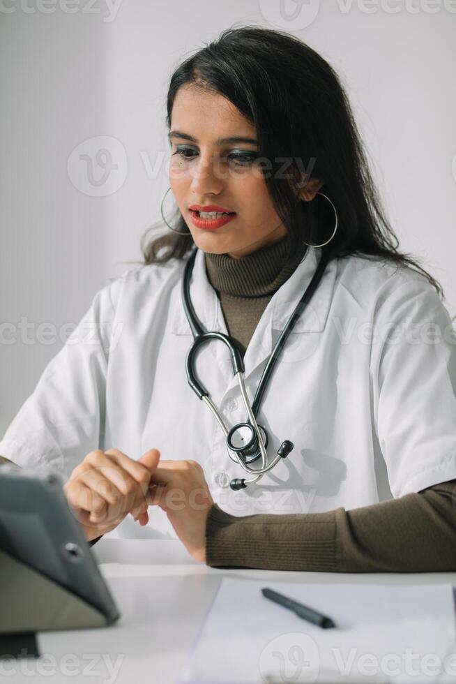 Indien femelle médecin dans une blanc manteau avec une stéthoscope conduit un en ligne consultation dans sa bureau. femelle médecin communique avec le téléspectateur directement dans le caméra photo