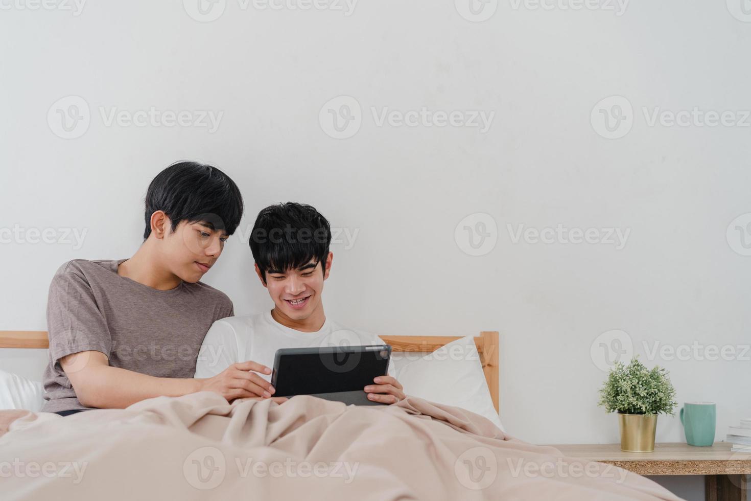 couple gay asiatique utilisant une tablette à la maison. jeunes hommes lgbtq asiatiques heureux de se détendre se reposer ensemble après le réveil, vérifier le courrier et les médias sociaux allongés sur le lit dans la chambre à la maison le matin concept. photo