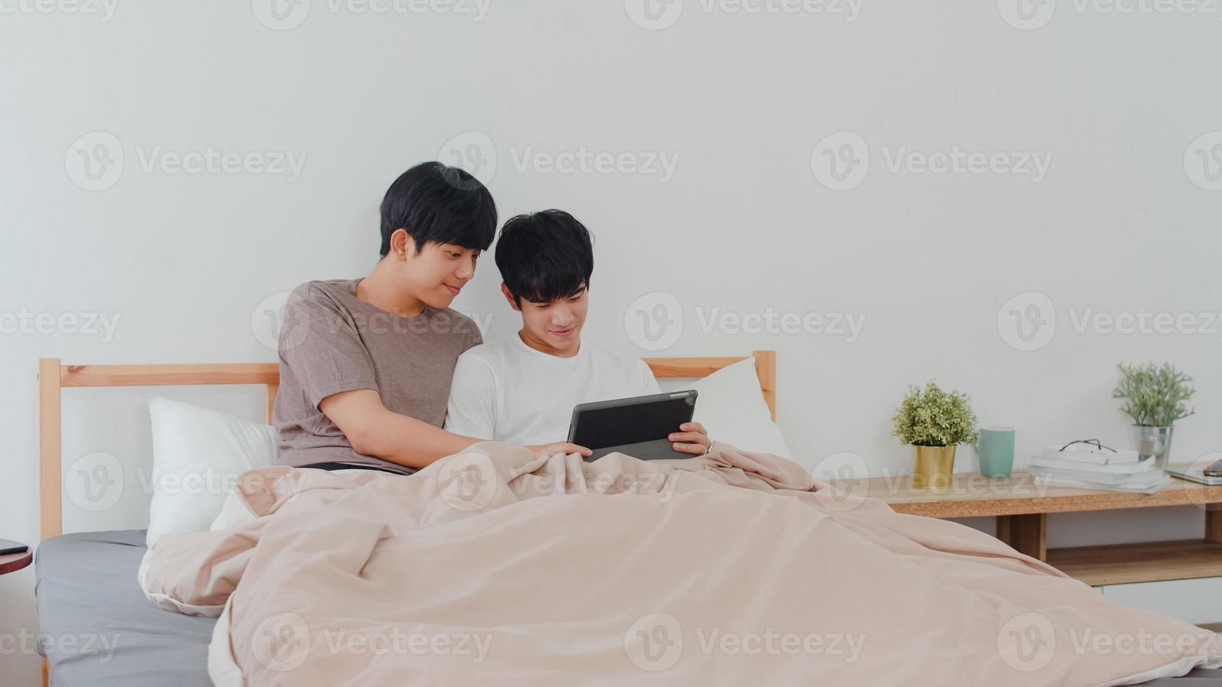 couple gay asiatique utilisant une tablette à la maison. jeunes hommes lgbtq asiatiques heureux de se détendre se reposer ensemble après le réveil, vérifier le courrier et les médias sociaux allongés sur le lit dans la chambre à la maison le matin concept. photo