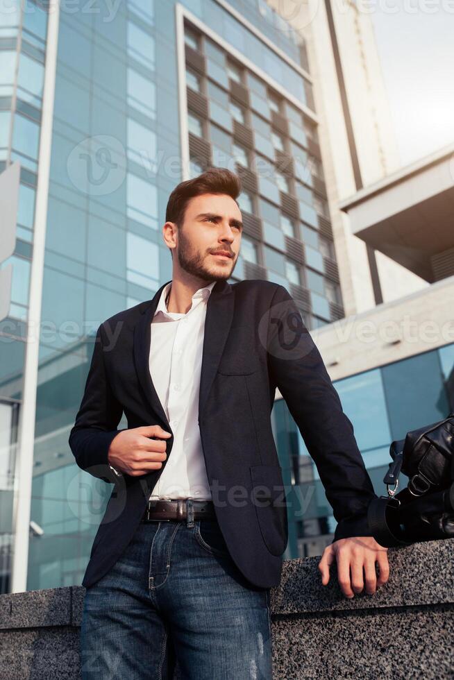 Beau Jeune homme d'affaire avec une barbe et dans une affaires costume permanent sur le rue contre le Contexte de le Bureau bâtiment suivant à une confortable élégant cuir sac. photo