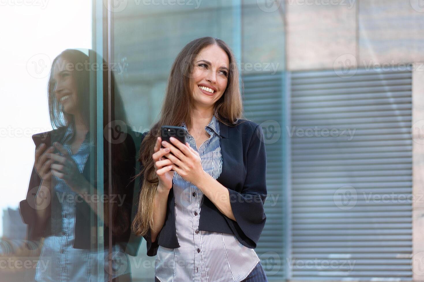 affaires femme 35 ans habillé Bande chemise et noir veste avec longue cheveux permanent près Bureau bâtiment Extérieur utilisation téléphone intelligent photo