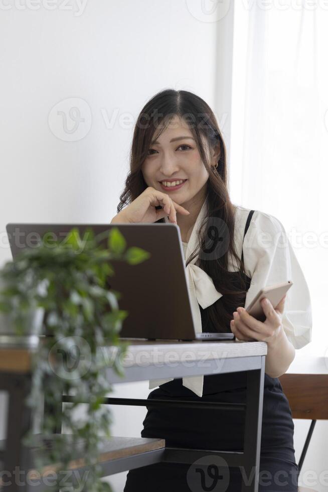 une Japonais femme vérification téléphone intelligent par éloigné travail dans le petit Bureau photo