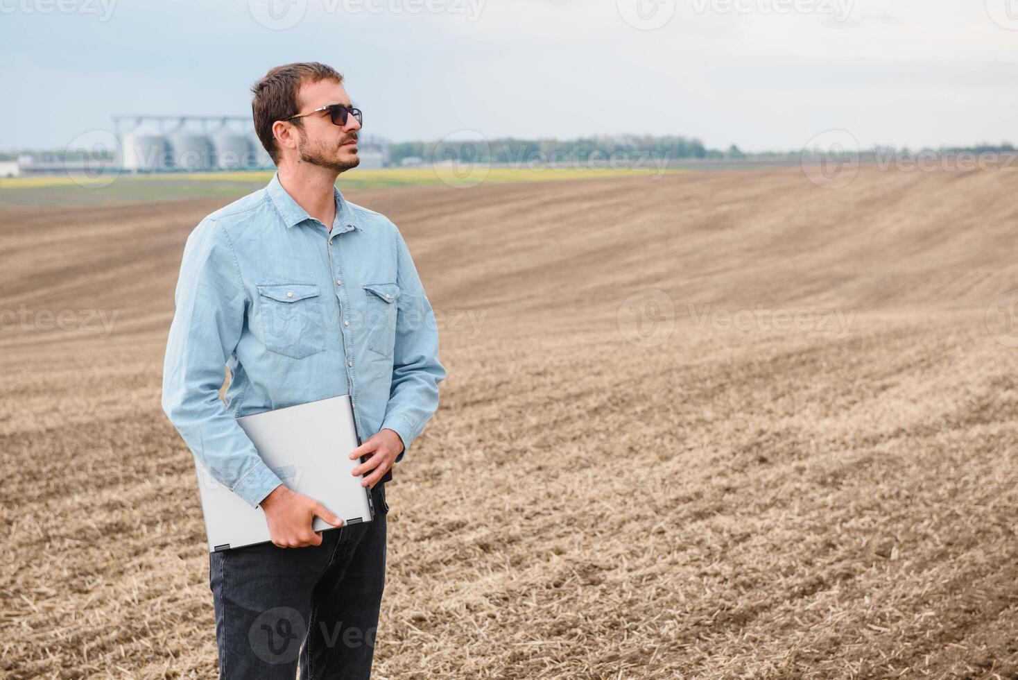 récolte concept. agriculteur dans une champ avec une portable sur une Contexte de une agricole silos pour espace de rangement et séchage de céréales, blé photo