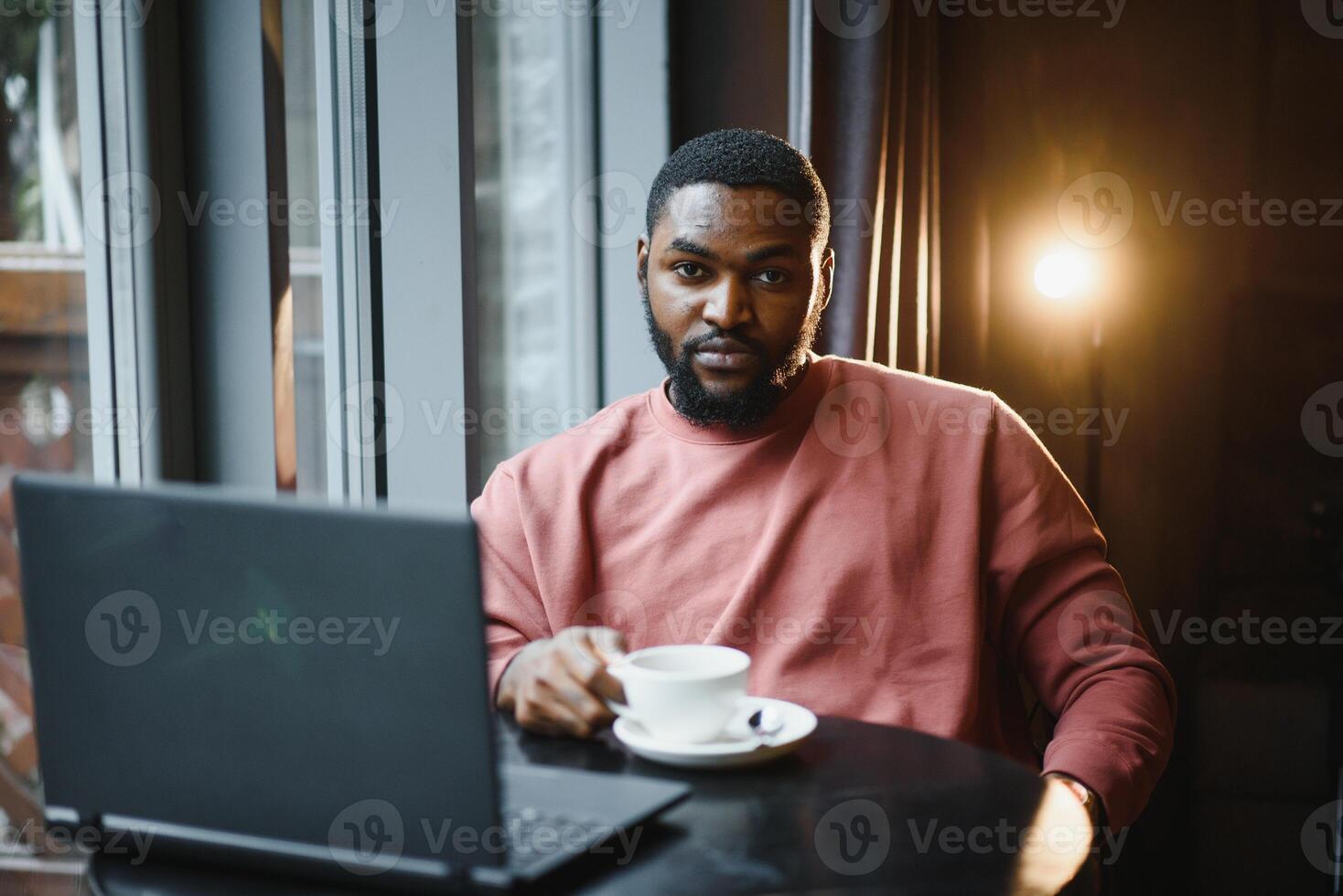 portrait de content africain homme d'affaire en utilisant téléphone tandis que travail sur portable dans restaurant. photo