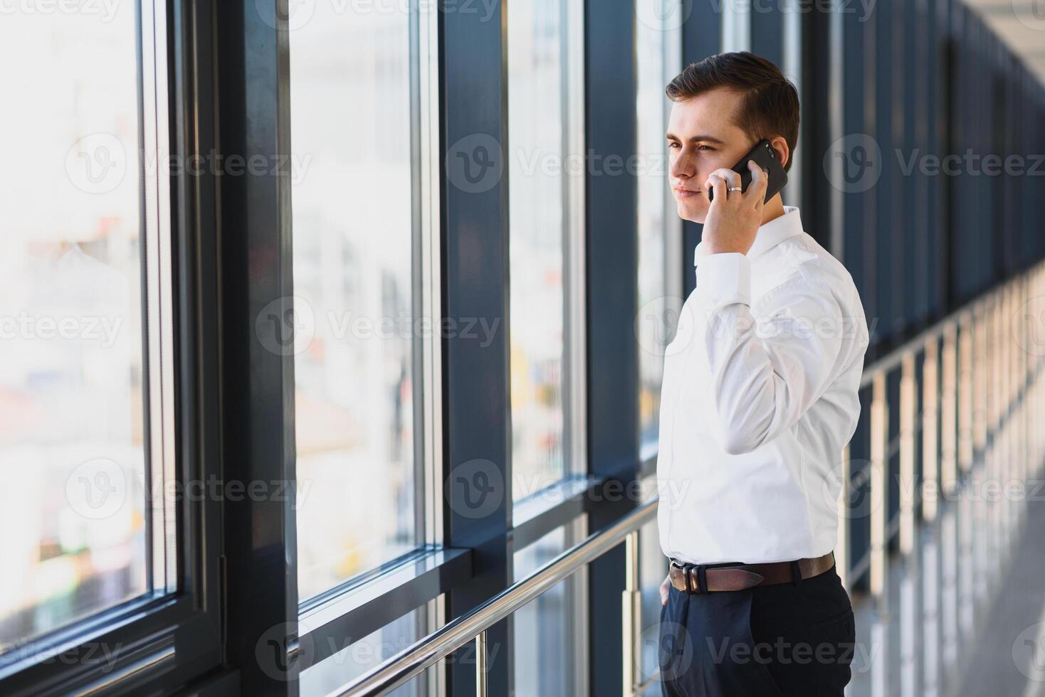 portrait de une sérieux sur de soi homme patron ayant mobile téléphone conversation tandis que repos après réunion avec le sien les partenaires, homme d'affaire parlant sur cellule Téléphone tandis que permanent dans moderne espace à l'intérieur photo