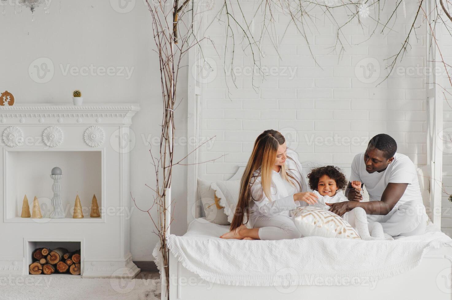 portrait de content multiracial Jeune famille mensonge sur confortable blanc lit à maison, souriant international maman et papa relaxant avec peu biracial fille enfant posant pour image dans chambre photo