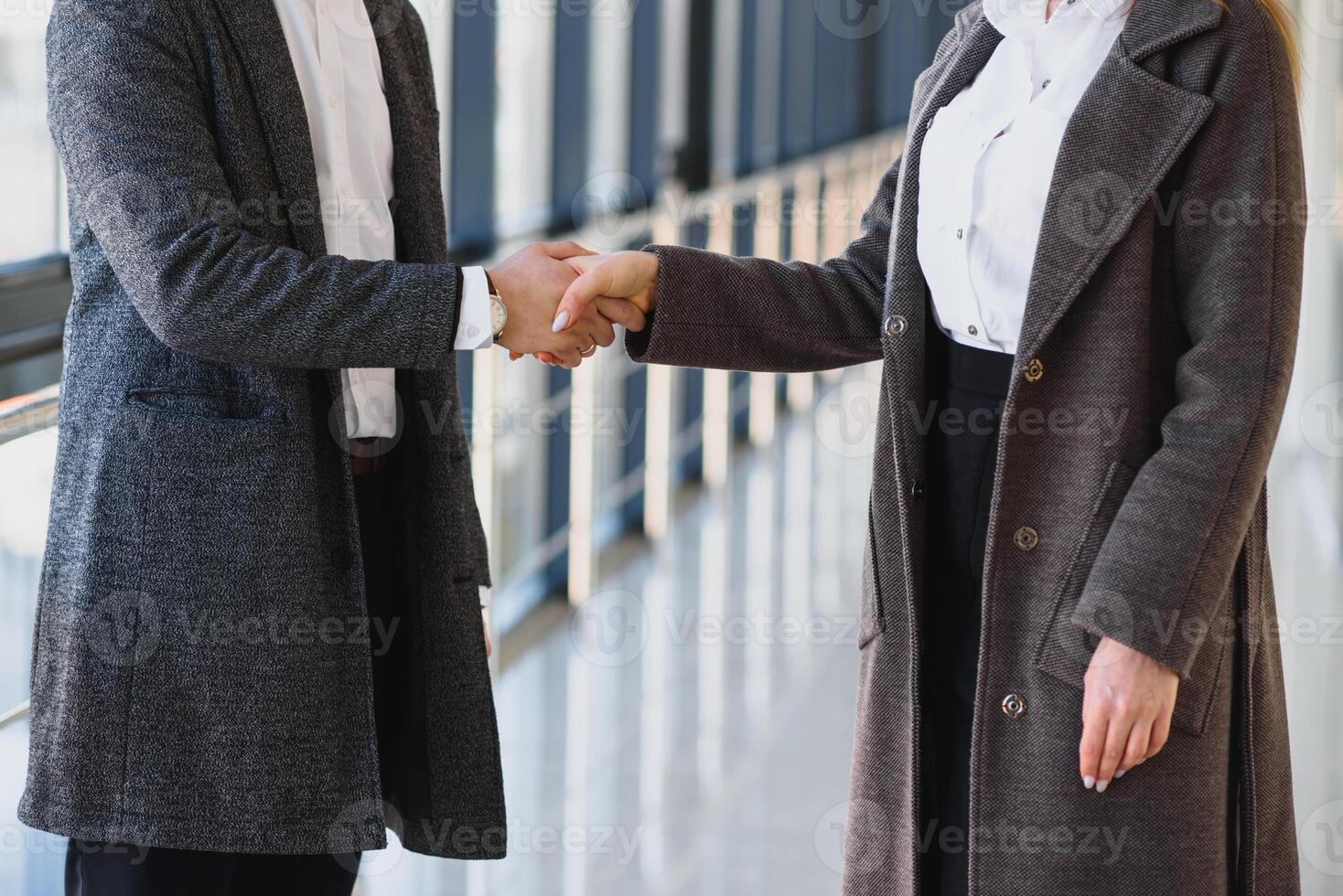 affaires Hommes et femmes tremblement mains avec une sourire sur le Contexte de le grand panoramique les fenêtres dans une moderne affaires centre. des modèles habillé dans une foncé affaires combinaisons. photo