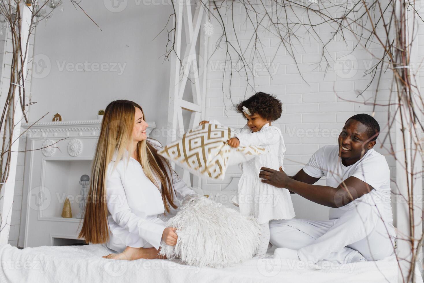 portrait de content multiracial Jeune famille mensonge sur confortable blanc lit à maison, souriant international maman et papa relaxant avec peu biracial fille enfant posant pour image dans chambre photo