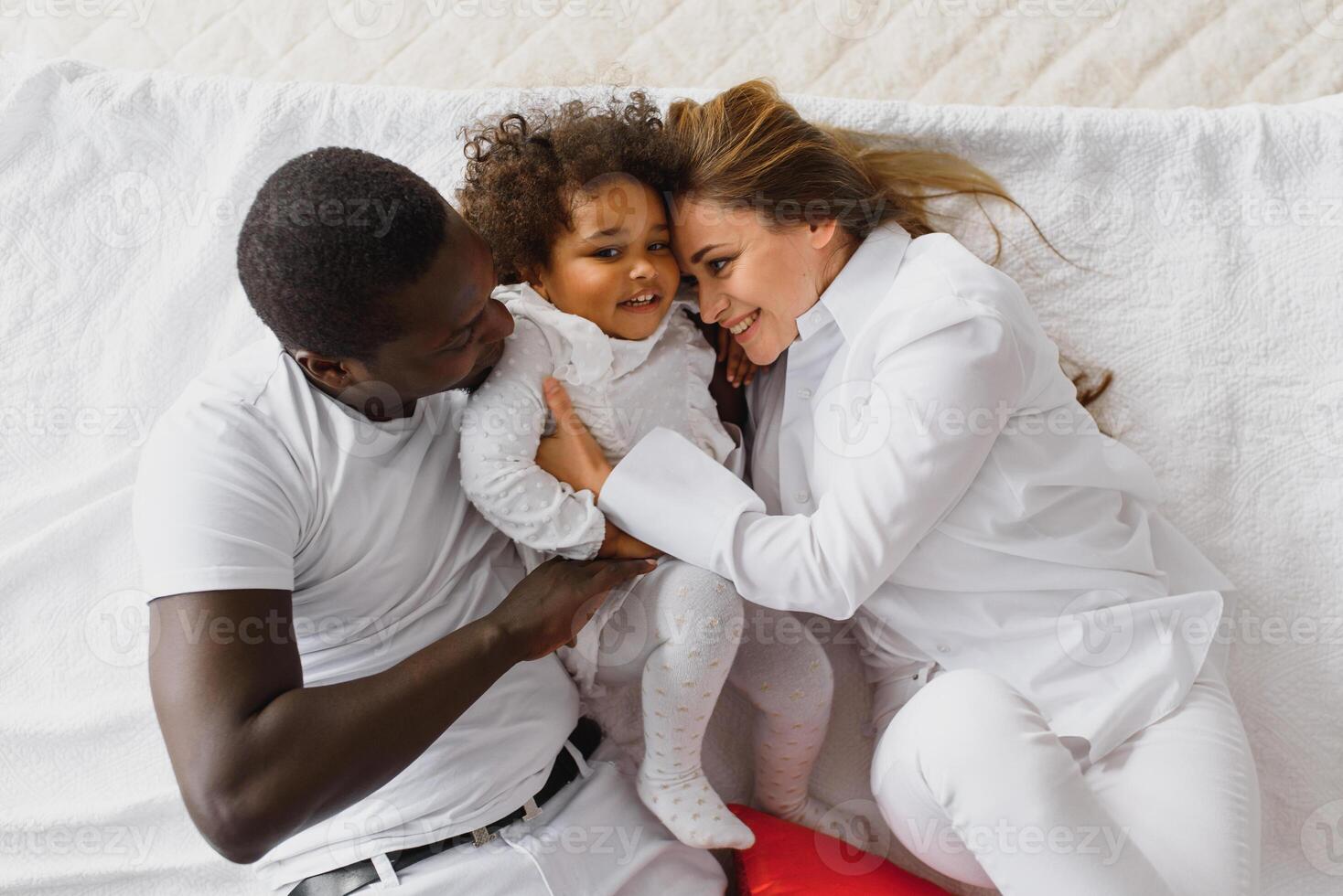 portrait de content multiracial Jeune famille mensonge sur confortable blanc lit à maison, souriant international maman et papa relaxant avec peu biracial fille enfant posant pour image dans chambre photo