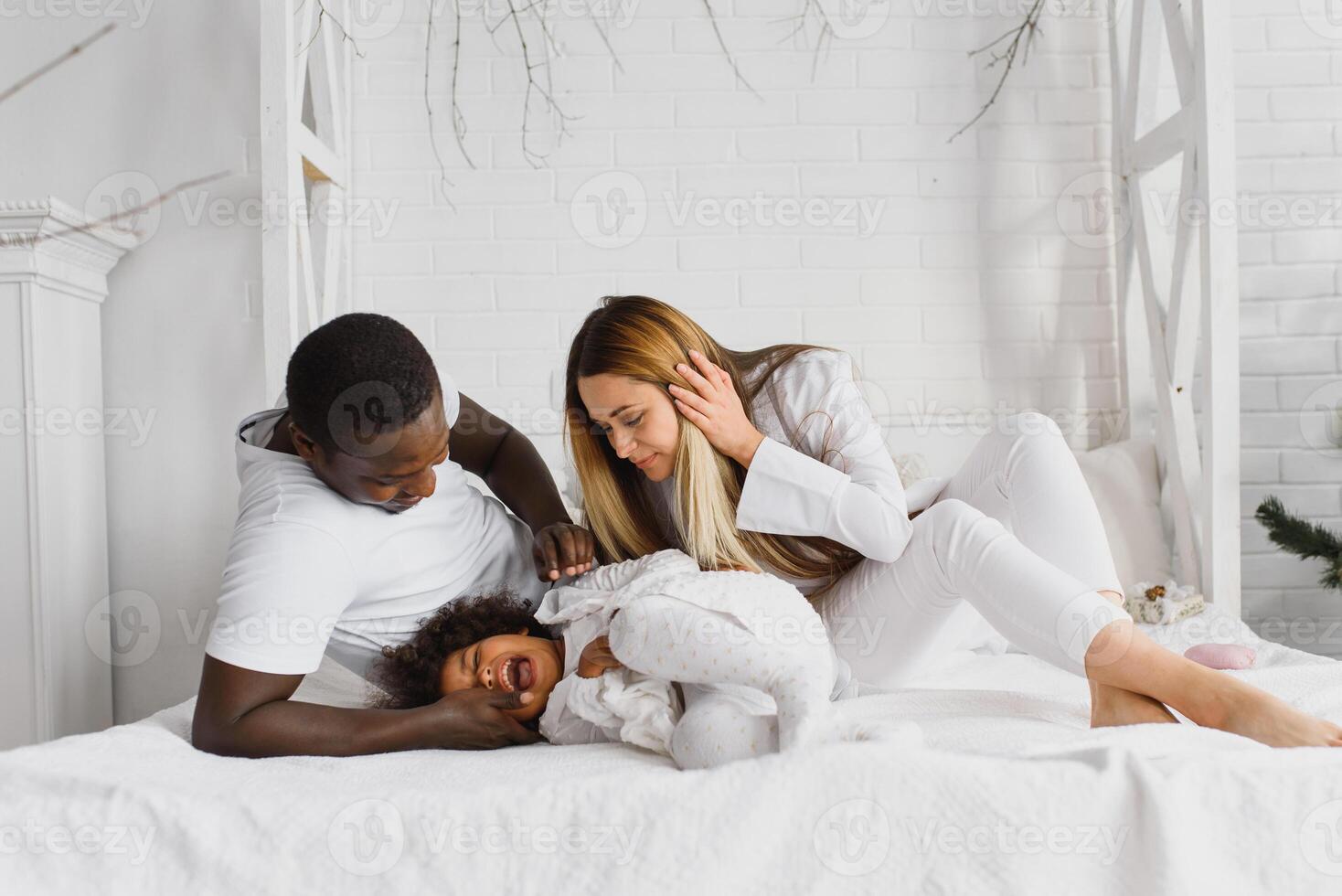 portrait de content multiracial Jeune famille mensonge sur confortable blanc lit à maison, souriant international maman et papa relaxant avec peu biracial fille enfant posant pour image dans chambre photo