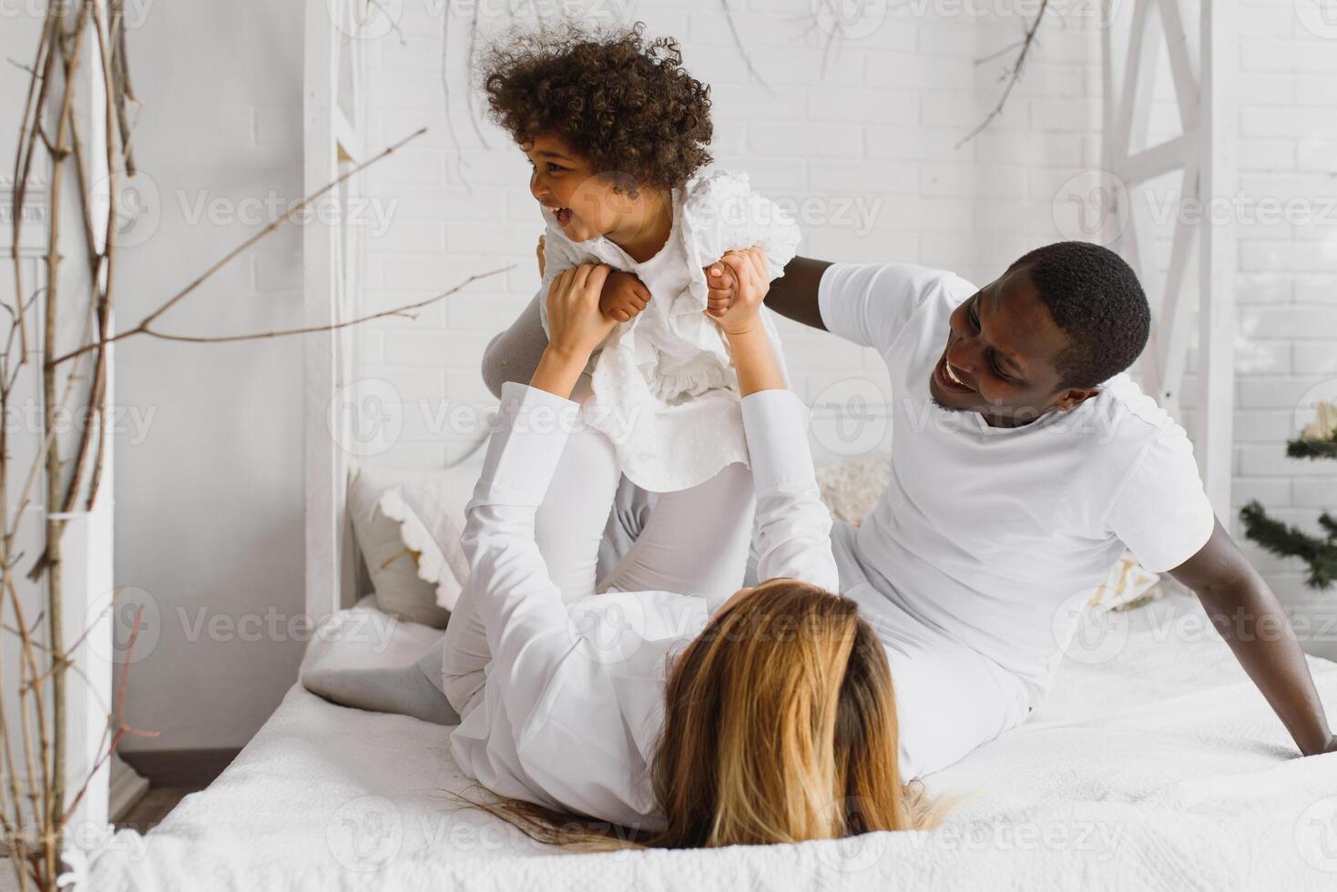 portrait de content multiracial Jeune famille mensonge sur confortable blanc lit à maison, souriant international maman et papa relaxant avec peu biracial fille enfant posant pour image dans chambre photo