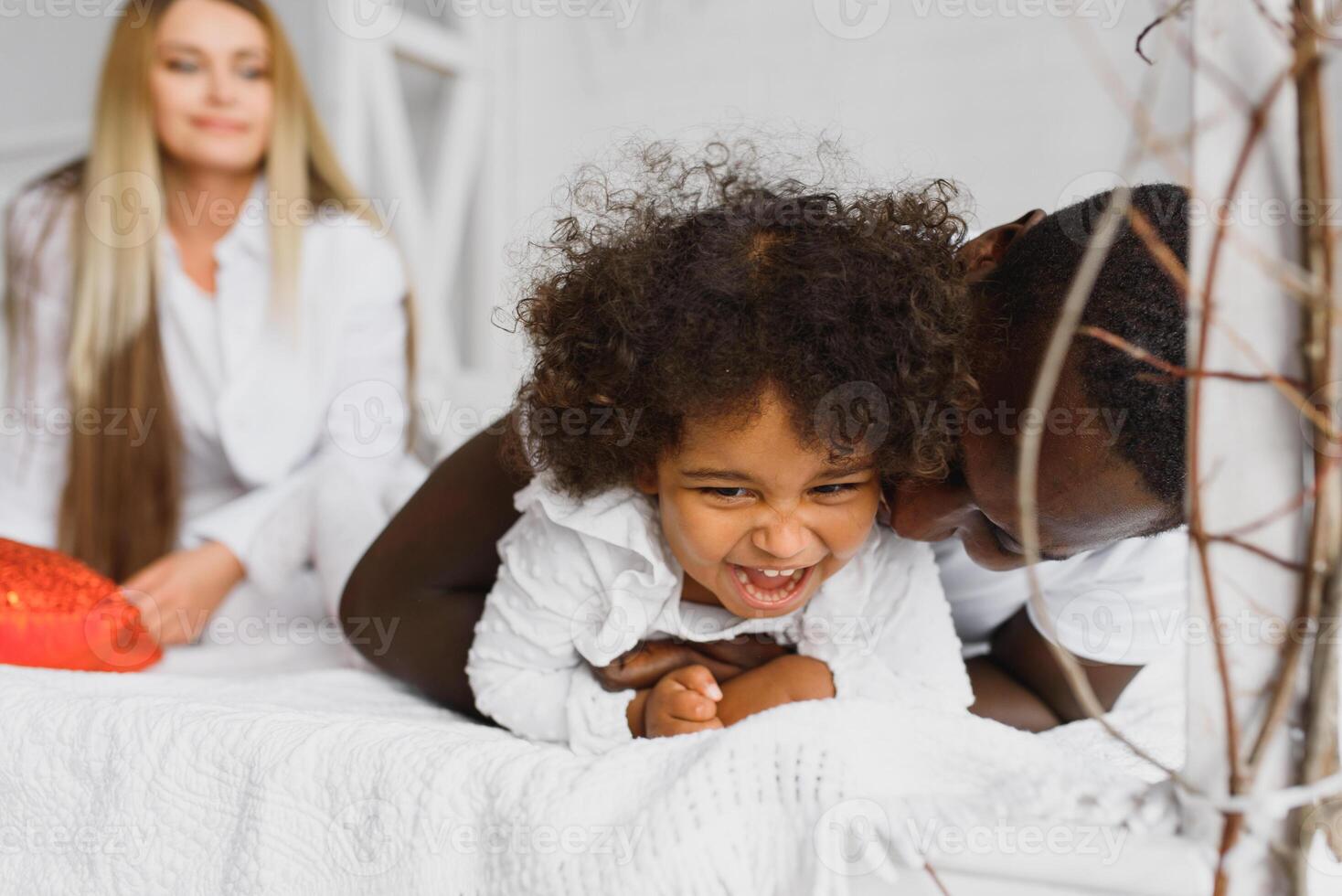 portrait de content multiracial Jeune famille mensonge sur confortable blanc lit à maison, souriant international maman et papa relaxant avec peu biracial fille enfant posant pour image dans chambre photo