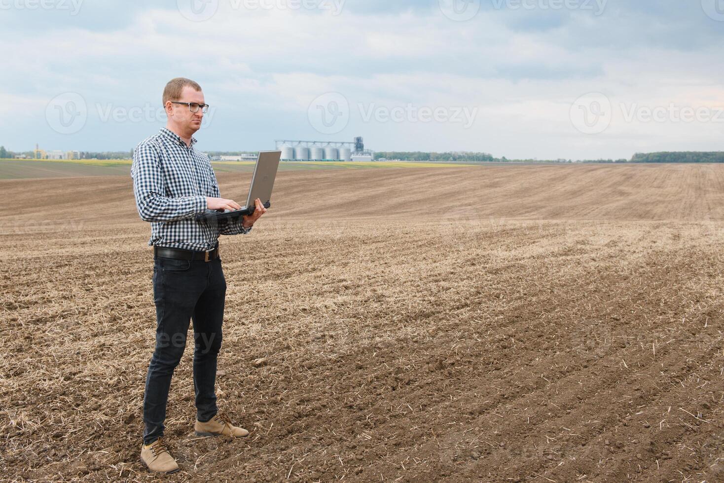 récolte concept. agriculteur dans une champ avec une portable sur une Contexte de une agricole silos pour espace de rangement et séchage de céréales, blé photo