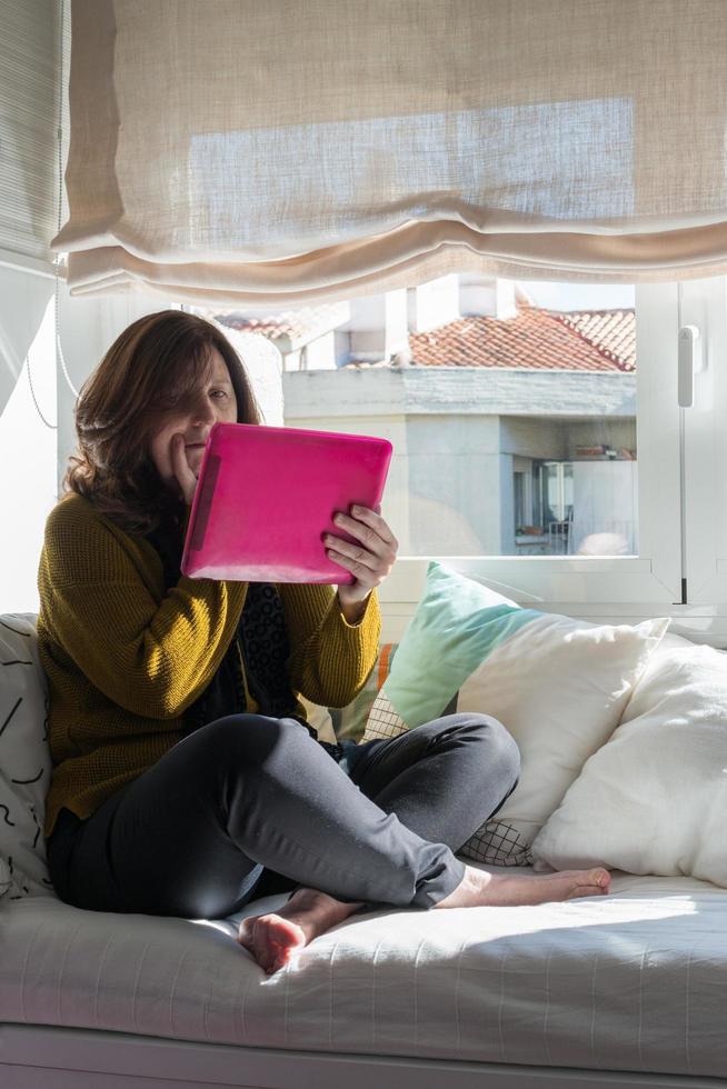 femme de race blanche âgée de 50 ans regardant une tablette avec inquiétude. assis de manière informelle sur un canapé près de la fenêtre. covid photo