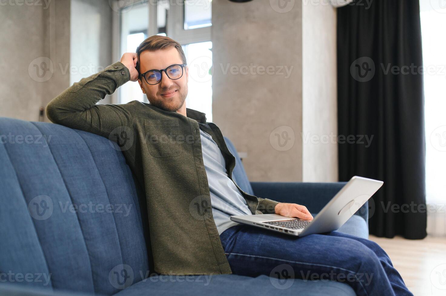 souriant Jeune homme portant des lunettes en utilisant ordinateur portable, séance dans confortable fauteuil, content Masculin à la recherche à ordinateur filtrer, bavardage dans social réseau ou achats en ligne, en jouant jeu, travail à maison. photo