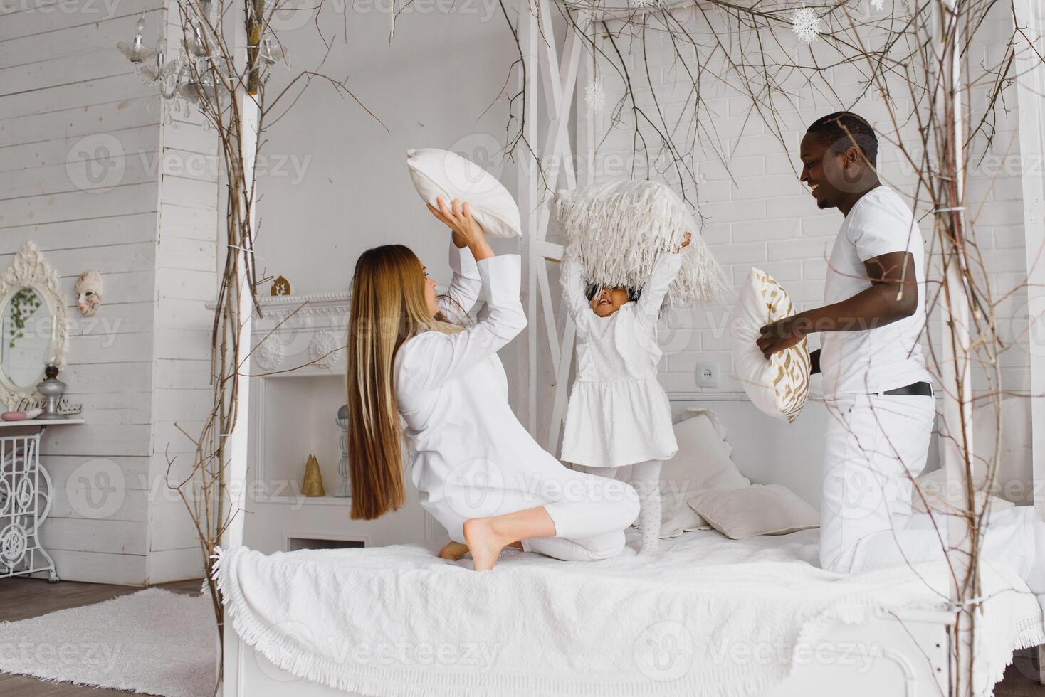 portrait de content multiracial Jeune famille mensonge sur confortable blanc lit à maison, souriant international maman et papa relaxant avec peu biracial fille enfant posant pour image dans chambre photo