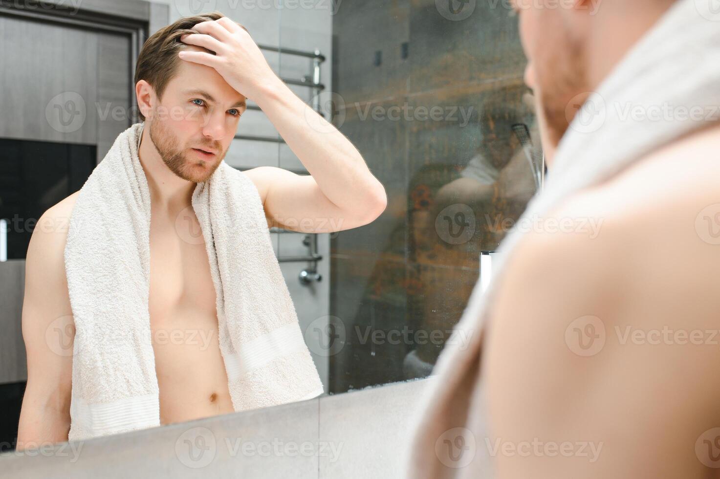 Jeune caucasien homme Regardez dans miroir faire soin de la peau faciale routine après douche dans Accueil salle de bains, millénaire Masculin dans serviette après une baignoire appliquer visage lotion ou crème sur peau après rasage, hygiène concept. photo