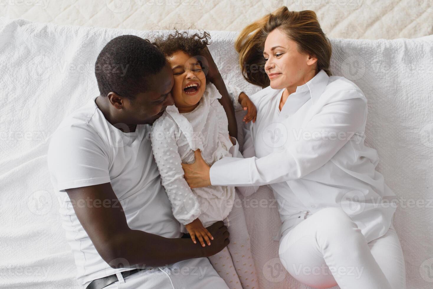 portrait de content multiracial Jeune famille mensonge sur confortable blanc lit à maison, souriant international maman et papa relaxant avec peu biracial fille enfant posant pour image dans chambre photo