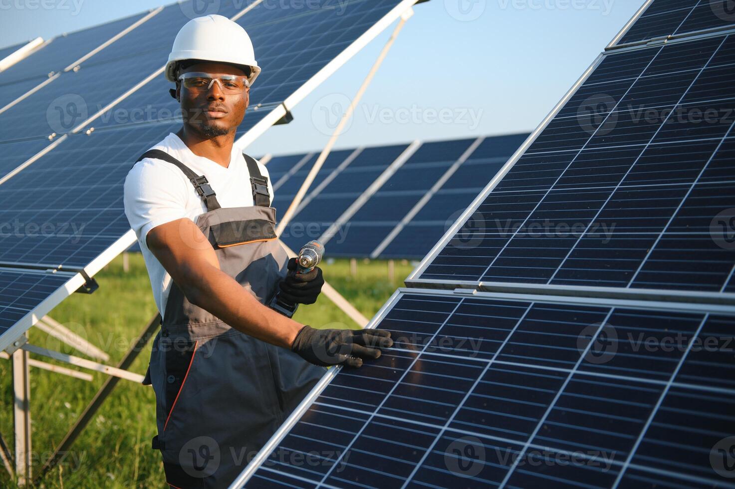 africain américain homme dans sécurité casque et des lunettes serrer des noisettes sur solaire panneaux avec Tournevis. compétent technicien en utilisant outils tandis que performant un service travail sur station photo