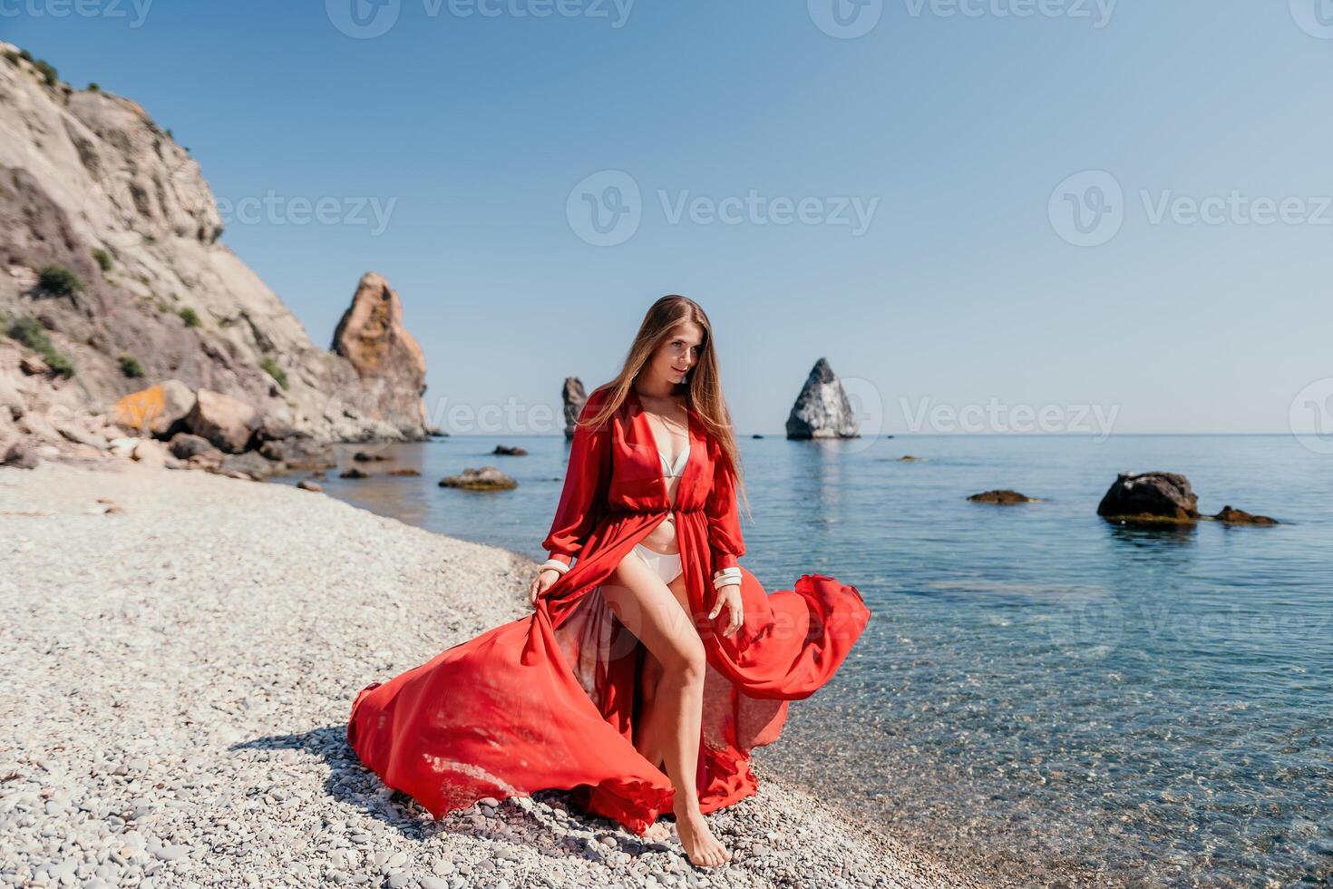 femme Voyage mer. content touristique dans rouge robe prendre plaisir prise image en plein air pour souvenirs. femme voyageur posant sur le Roche à mer baie entouré par volcanique montagnes, partage Voyage aventure périple photo