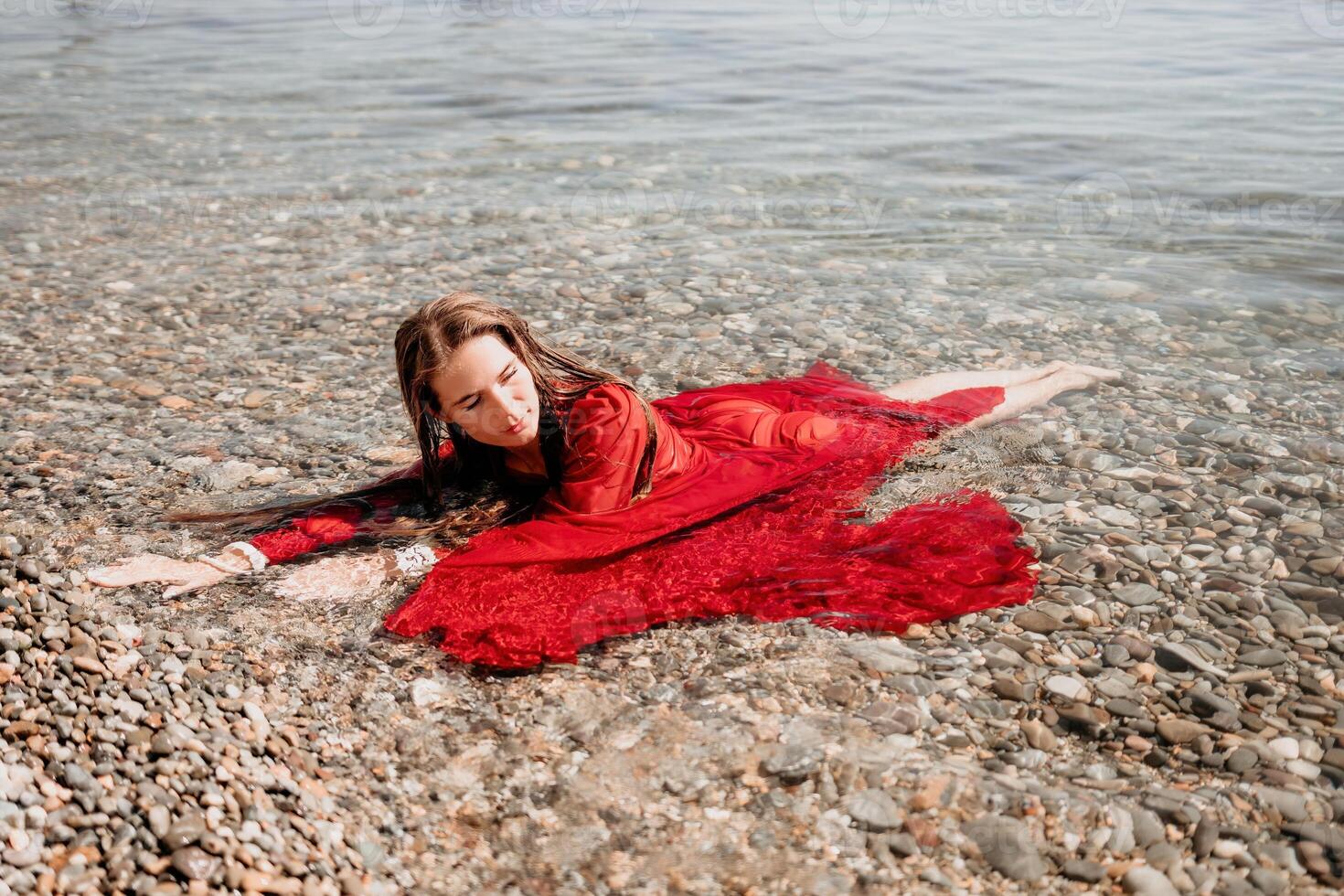 femme Voyage mer. content touristique dans rouge robe prendre plaisir prise image en plein air pour souvenirs. femme voyageur posant dans mer plage, entouré par volcanique montagnes, partage Voyage aventure périple photo