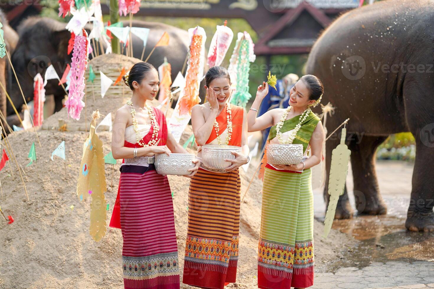 magnifique thaïlandais Jeune Dame vaisselle thaïlandais traditionnel robe utilisation fleurs à saupoudrer l'eau sur chaque autre sur le thaïlandais Nouveau années journée dans une amusement façon sur flou l'éléphant et pile de le sable Contexte. photo