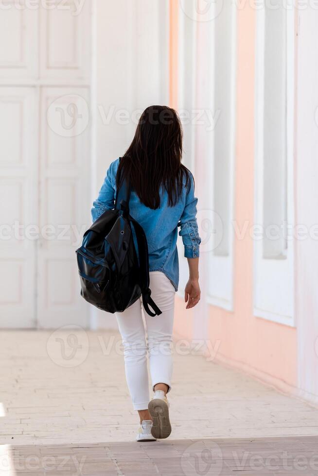 femme avec longue marron cheveux en marchant avec sac à dos. vue de le dos. blanc jeans et bleu chemise. lumière salle Contexte. photo
