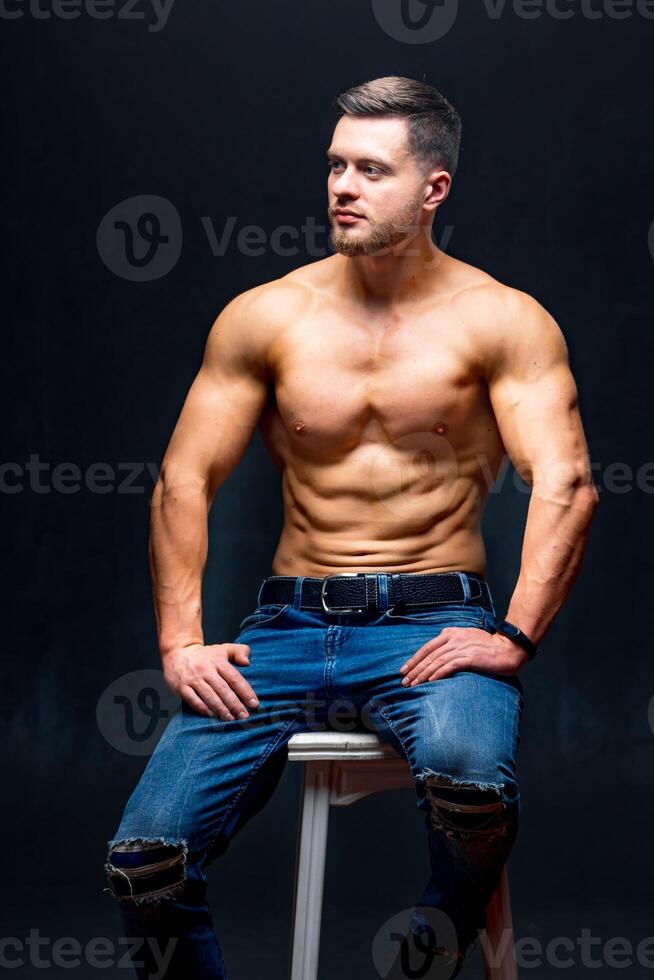 Beau fort gars homme sans pour autant une chemise contre foncé Contexte. séance sur chaise. studio photo. Masculin beauté concept. fermer. photo